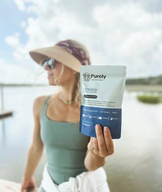 A woman relaxing outdoors by the water, holding a pouch of Purely Mushroom Unwind gummies